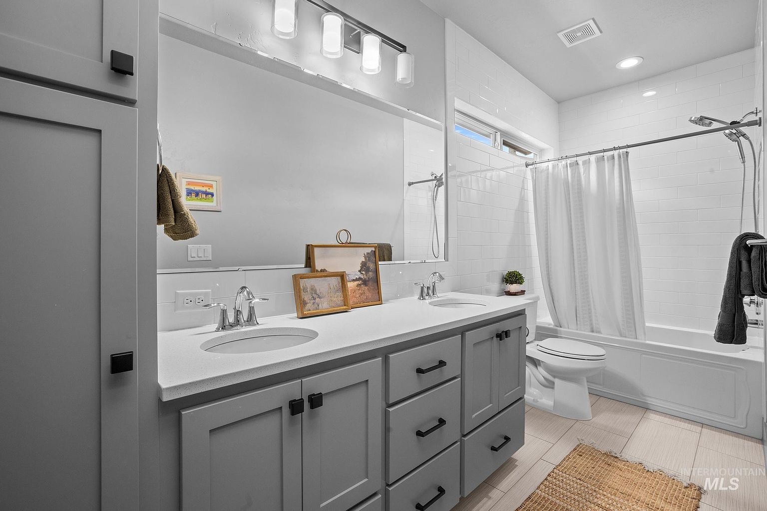 This bright and modern bathroom features a dual-sink vanity with light gray cabinetry and a white countertop. A large, wide mirror spans the wall above the vanity, complemented by a contemporary light fixture. To the right, a shower-tub combination is enclosed by a white curtain and features white subway-style wall tiles, creating a clean and cohesive aesthetic.