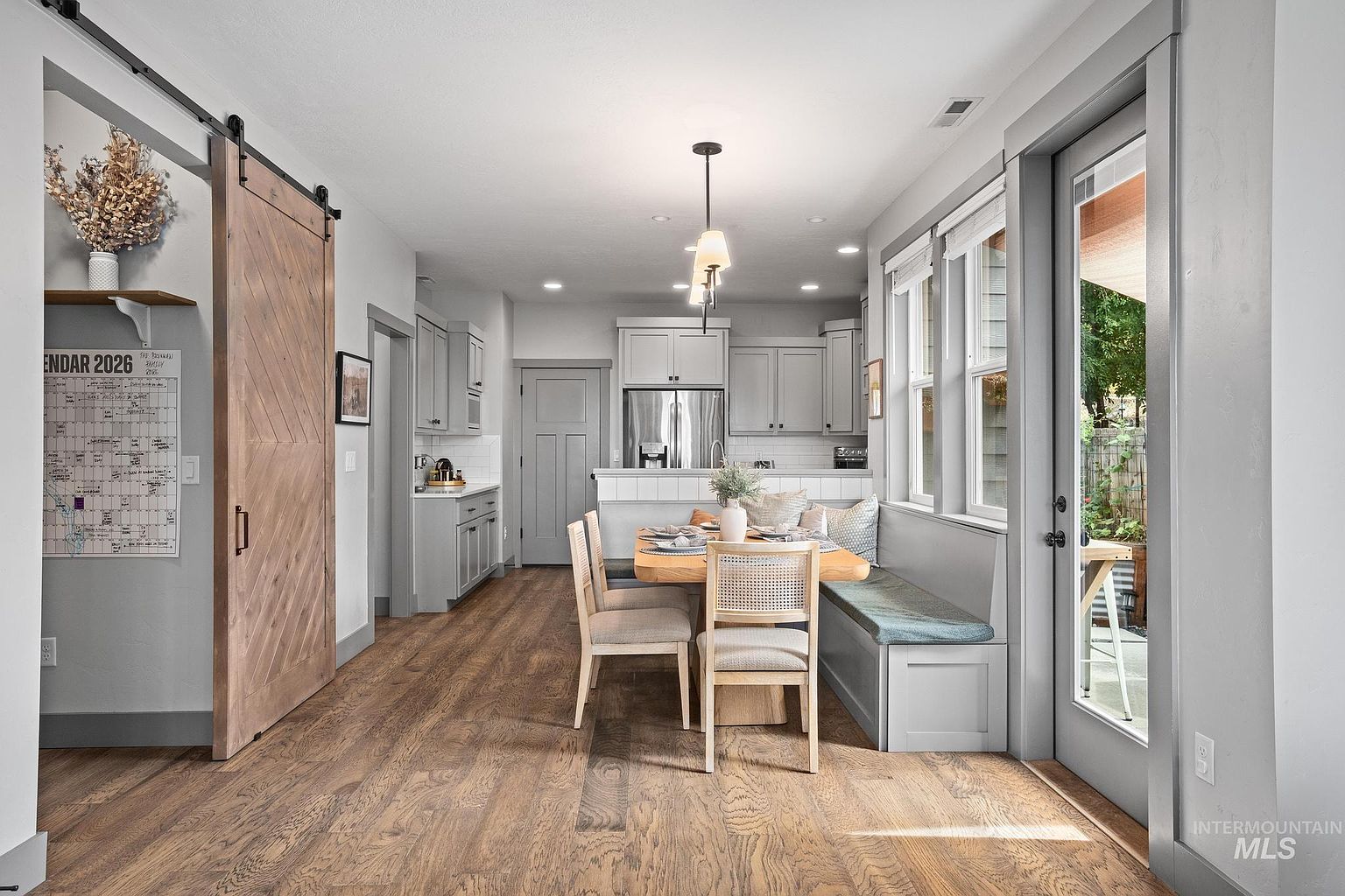 This bright dining area features a built-in bench seating arrangement paired with a light wood table and two cane-back chairs. The space is anchored by warm-toned wood flooring and illuminated by a modern pendant light, while a large sliding barn door adds a rustic touch to the side wall. Large windows and a glass-paneled door provide ample natural light and a view of the outdoor space, creating an inviting and functional breakfast nook.