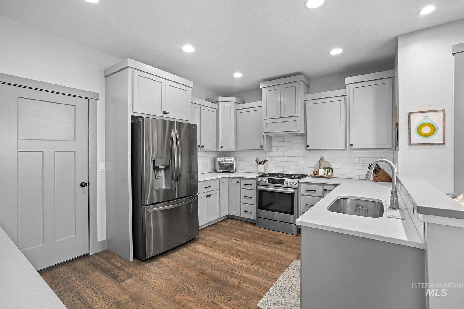 This modern kitchen features light gray cabinetry, stainless steel appliances, and a white subway tile backsplash. The space includes a peninsula with an undermount sink and light-toned wood flooring, creating a bright and functional layout. The perspective is a wide-angle shot from the kitchen entrance, showcasing the efficient work triangle and clean, contemporary aesthetic.