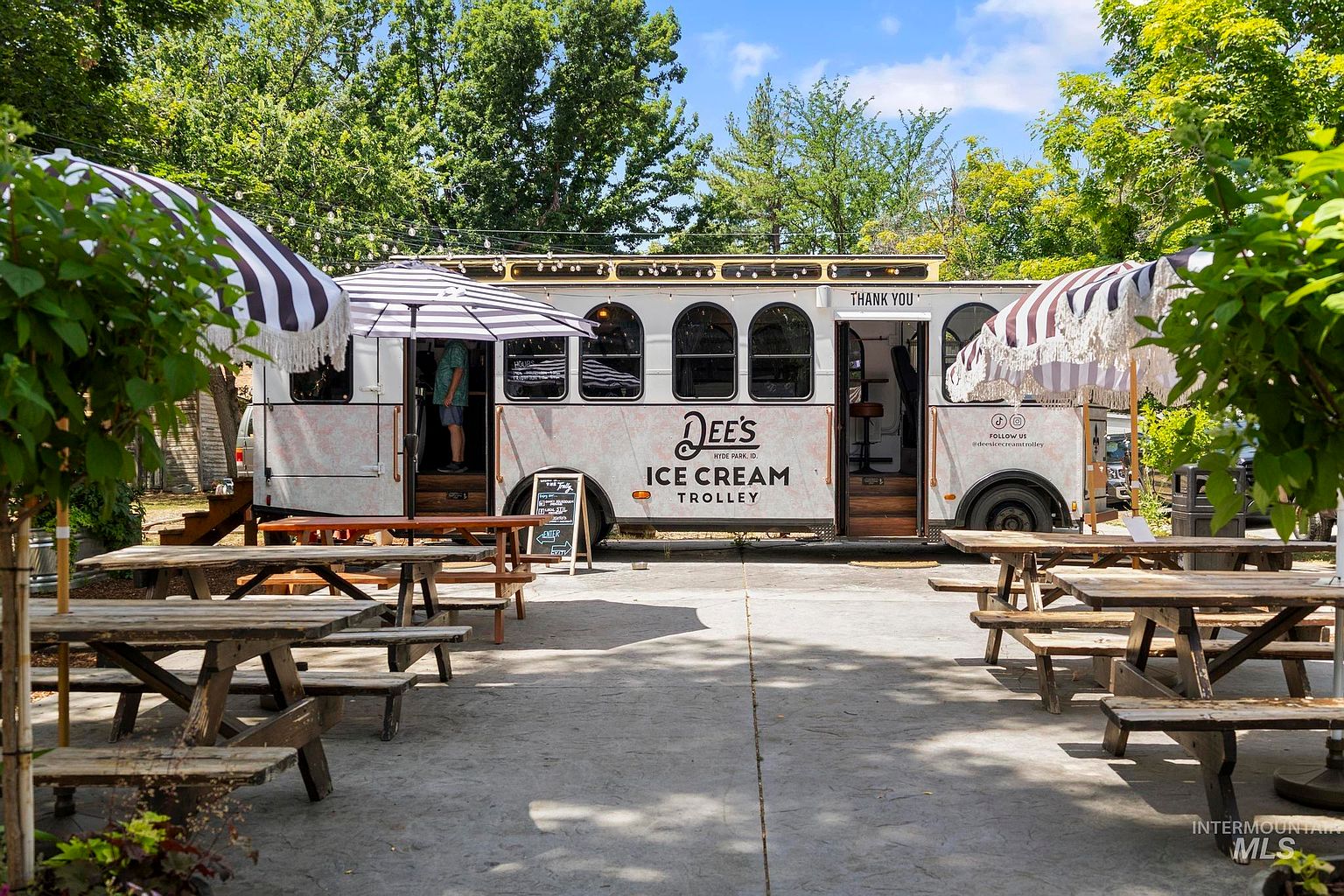 This image captures a charming outdoor seating area featuring a converted trolley serving as an ice cream shop named 'Dee's Ice Cream Trolley.' The scene is set on a paved patio with multiple wooden picnic tables shaded by black-and-white striped umbrellas, creating a welcoming and nostalgic atmosphere. Lush green trees provide a natural canopy overhead, enhancing the relaxed, community-focused vibe of this unique local amenity.