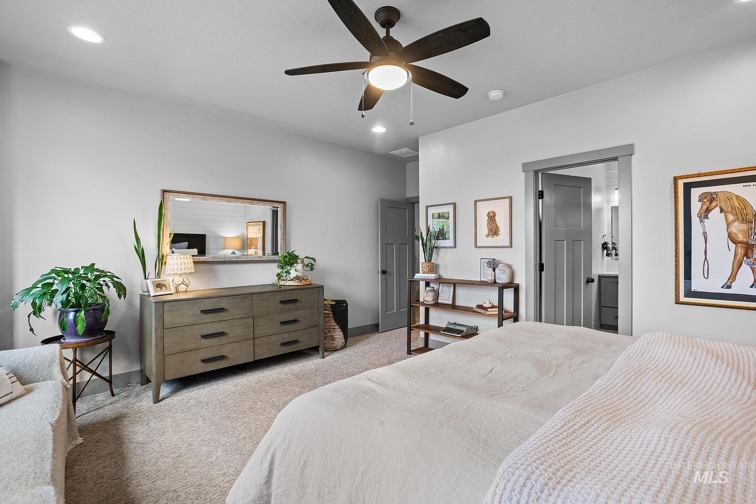 This primary bedroom features a neutral color palette with soft gray walls and plush carpeting, creating a serene and inviting atmosphere. The room is anchored by a large bed with light-toned bedding, complemented by a wooden dresser topped with a mirror, a decorative plant, and a small lamp. A wooden shelving unit stands near the doorway, which leads into an en-suite bathroom, while a framed horse print adds a touch of character to the wall.