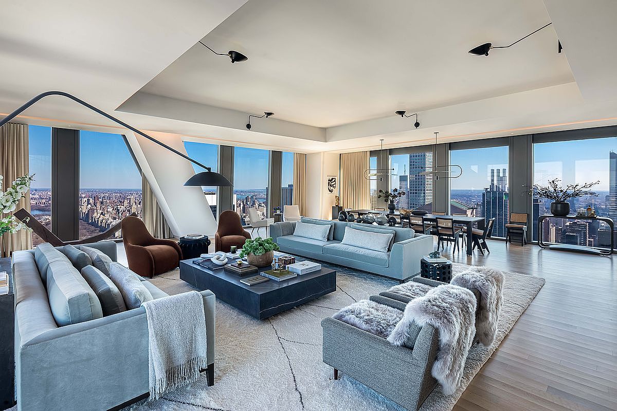 This expansive, high-rise living room features a sophisticated open-concept layout with floor-to-ceiling windows that provide panoramic city views. The space is furnished with two plush sofas, a large central stone coffee table, and two accent chairs, all arranged on a textured area rug. A dining area is visible in the background, and the room is illuminated by minimalist ceiling-mounted lighting fixtures, creating a luxurious and airy atmosphere.