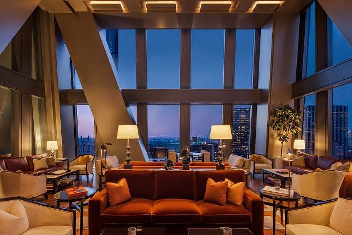 This sophisticated high-rise living room features a symmetrical layout centered around a plush, rust-colored velvet sofa. The space is defined by expansive floor-to-ceiling windows that offer a panoramic view of the city skyline at dusk, framed by structural columns. Elegant table lamps provide warm ambient lighting, complementing the refined, contemporary furniture arrangement that creates an inviting atmosphere for entertaining.