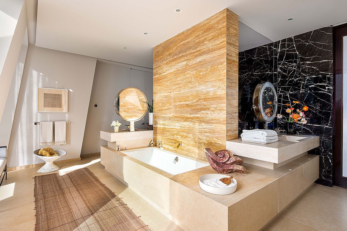 This luxurious primary bathroom features a central soaking tub set against a striking, tall stone-clad wall with warm, horizontal veining. The space is anchored by dual vanity areas with stone countertops and circular mirrors, contrasted by a dramatic black stone wall on the right. A woven rug adds texture to the light-toned stone flooring, creating a sophisticated and serene spa-like atmosphere.
