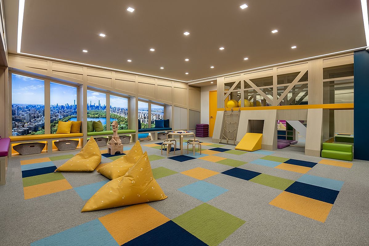 This vibrant children's playroom features a colorful, modular carpet design and multiple beanbag chairs, creating a playful and inviting atmosphere. The space includes a built-in climbing structure with a slide and a rope ladder, alongside a small table and chairs for activities. Large windows along the back wall display a panoramic view of a city skyline, adding a unique urban backdrop to the indoor play area.
