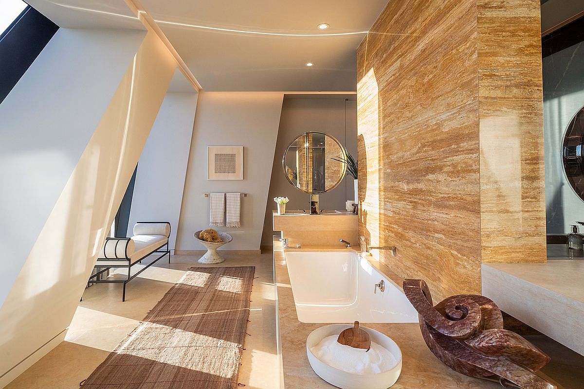 This luxurious primary bathroom features a striking wall clad in warm-toned, horizontally-veined stone that serves as a dramatic backdrop for the deep soaking tub. The space is bathed in natural light from large, angled windows, highlighting the minimalist vanity with a circular mirror and a sleek bench seat. A sculptural, dark-toned decorative piece sits near the tub, adding an artistic touch to the sophisticated, spa-like atmosphere.