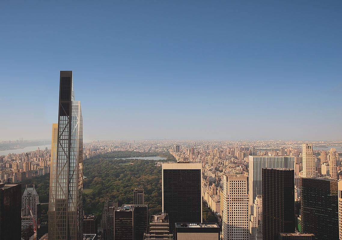 This high-altitude aerial perspective captures a sweeping view of the New York City skyline, dominated by the prominent, modern glass skyscraper in the foreground. Central Park stretches out as a vast green expanse in the middle ground, surrounded by a dense urban landscape of high-rise buildings. The clear blue sky provides a bright, expansive backdrop that emphasizes the scale and density of the metropolitan environment.