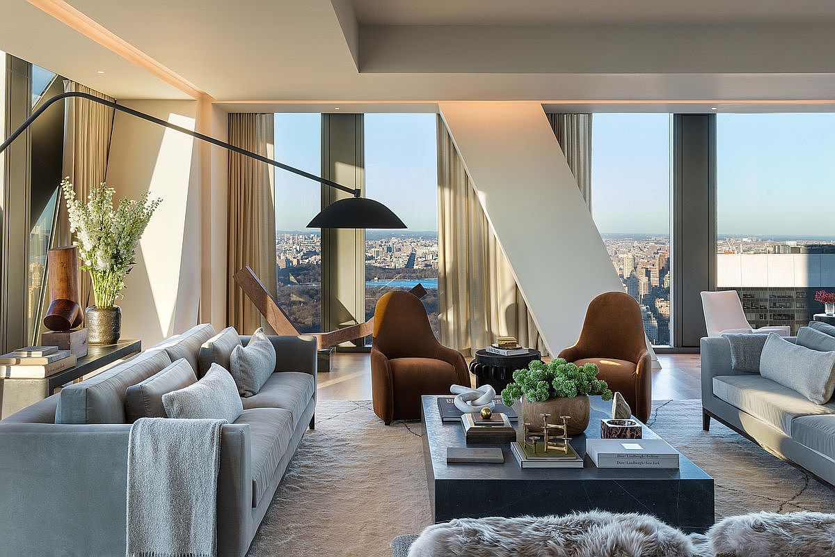 This sophisticated, high-rise living room features a contemporary design with two plush grey sofas and a pair of warm-toned accent chairs arranged around a large, dark stone coffee table. The space is defined by expansive floor-to-ceiling windows that offer a panoramic view of the city skyline and a central park. A dramatic, oversized arched floor lamp anchors the seating area, while minimalist decor and soft textures create an atmosphere of refined luxury.