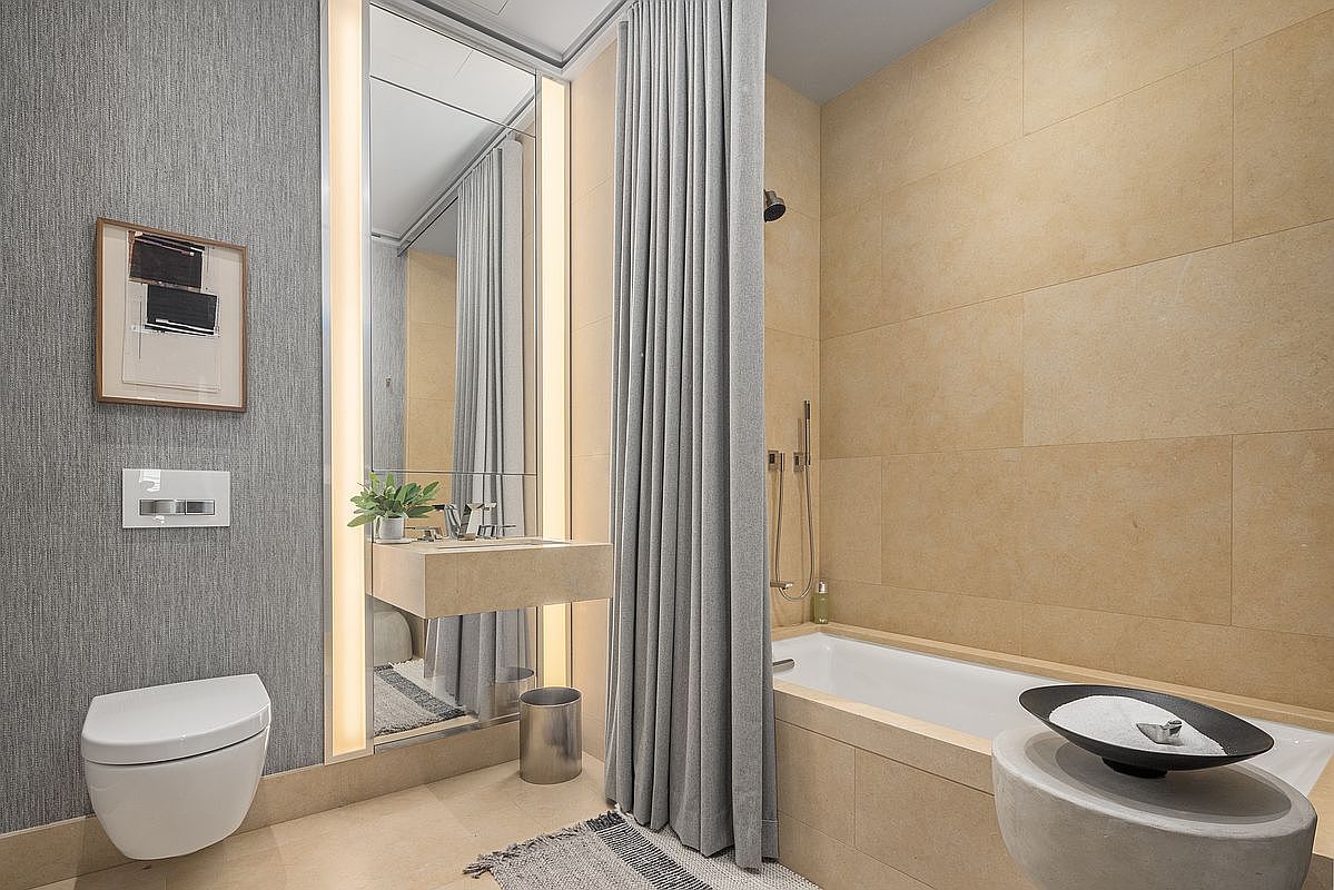 This contemporary bathroom features a wall-mounted toilet, a floating stone vanity, and a bathtub enclosed by a floor-to-ceiling grey curtain. The walls are clad in large, neutral-toned stone tiles, complemented by a textured grey wallpaper on the adjacent wall. A tall, illuminated mirror adds a sleek, modern touch to the space, creating a sophisticated and minimalist aesthetic.