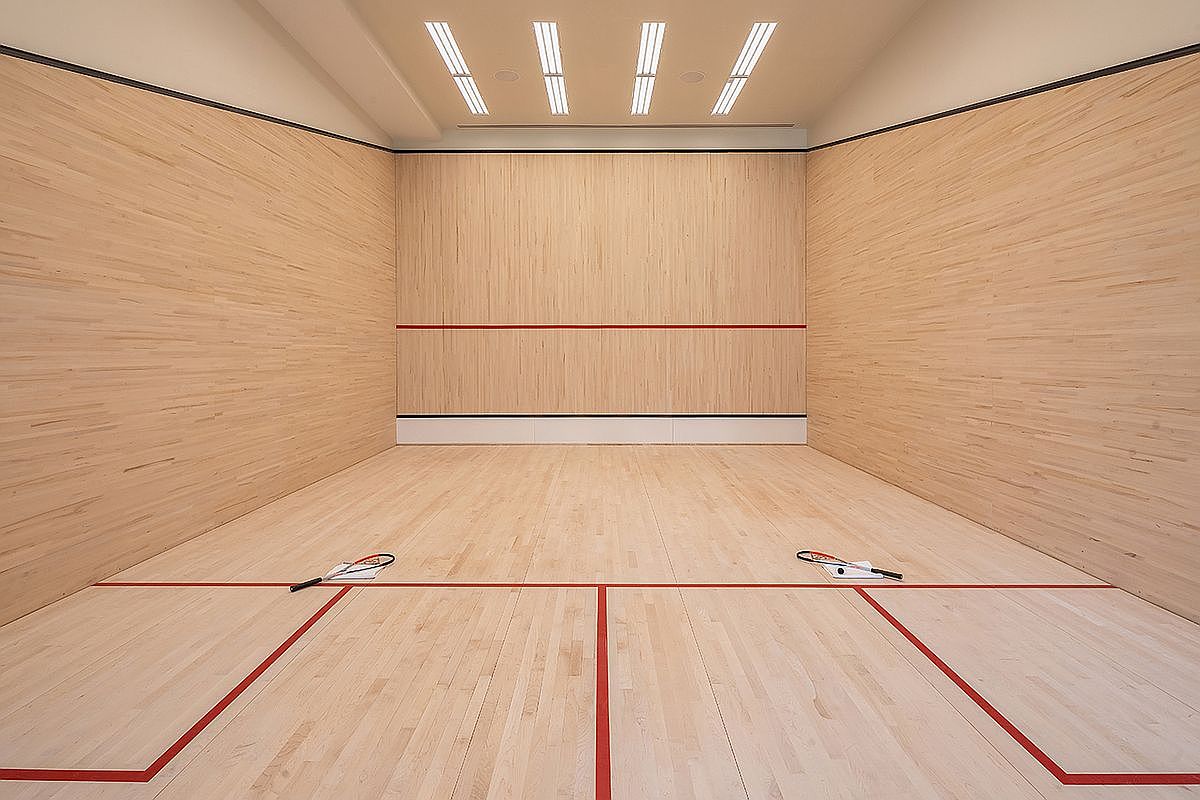 This image captures a professional-grade squash court featuring light wood paneling on all walls and a matching hardwood floor. The court is marked with crisp red lines for gameplay and includes two rackets resting on the floor, emphasizing its functional purpose. The perspective is centered and symmetrical, looking down the length of the court toward the front wall, which is illuminated by recessed ceiling lighting.