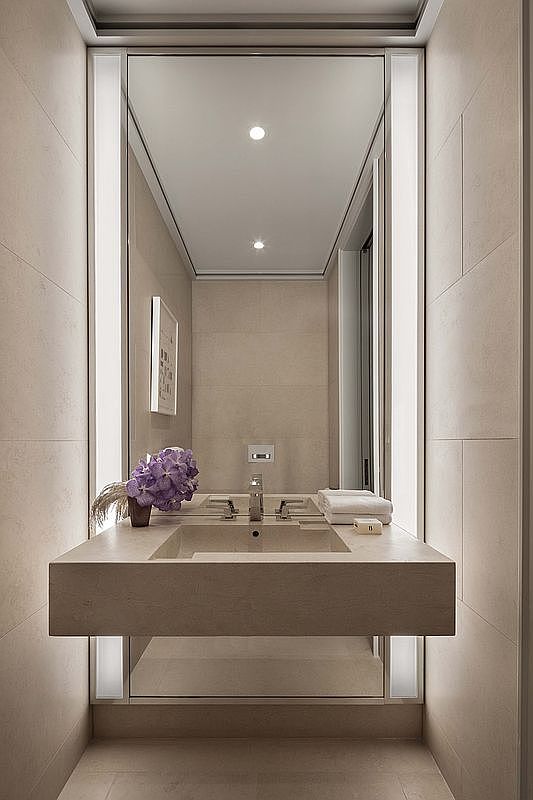 This minimalist powder room features a floating stone vanity with a sleek, integrated sink and modern chrome fixtures. A large, full-height mirror is flanked by vertical integrated light strips, creating a bright and sophisticated atmosphere. The space is finished with neutral-toned stone wall tiles and includes a small vase of purple flowers for a touch of color.