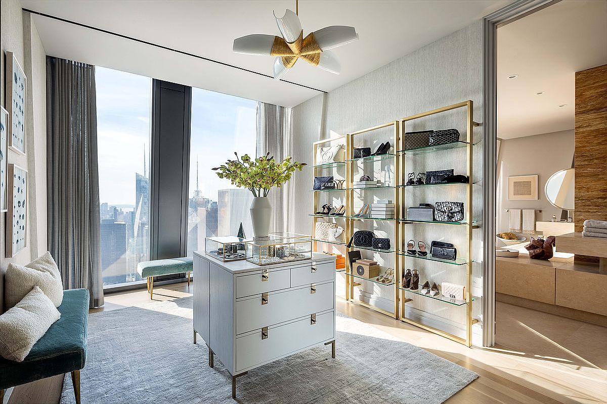This sophisticated walk-in closet features a central white dresser topped with glass display cases and a vibrant floral arrangement, set against a backdrop of floor-to-ceiling windows offering expansive city views. The space is organized with gold-toned open shelving units displaying luxury handbags and accessories, while a plush velvet bench provides seating. A doorway leads into an adjacent bathroom, creating a seamless flow between the dressing area and personal care space.