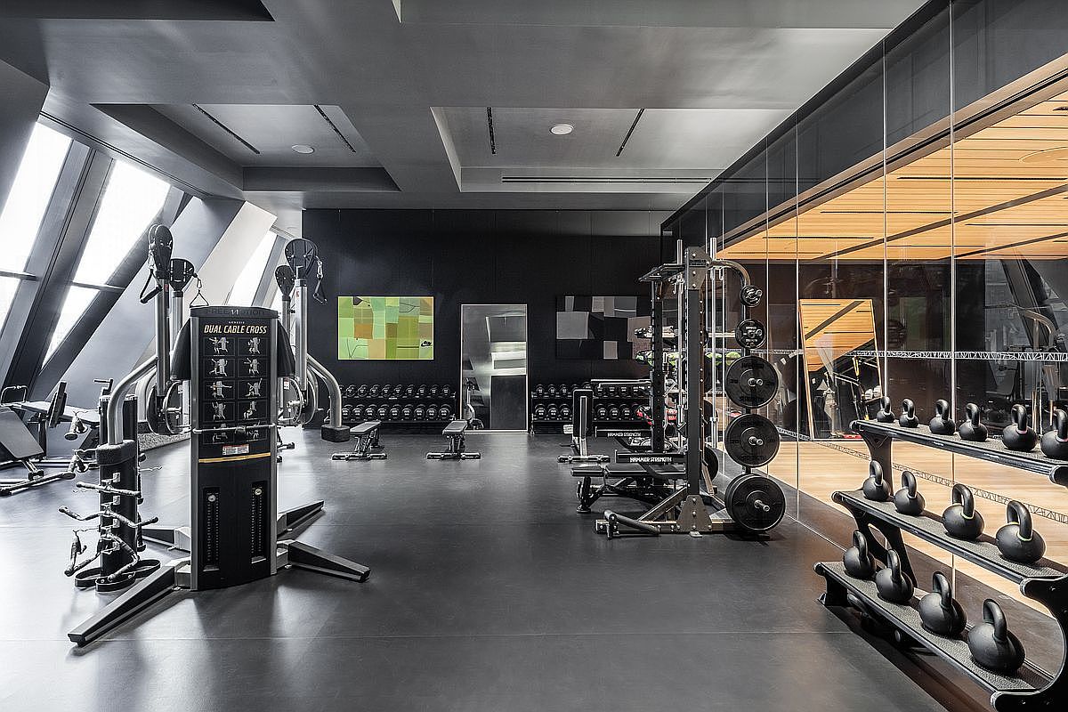 This sleek, modern home gym features a dark, minimalist aesthetic with high-end fitness equipment, including a dual cable cross machine and a weight rack. The space is defined by a dark color palette, smooth flooring, and a glass-walled section that reveals a warm, wood-toned area beyond. Large, angled windows on the left allow natural light to flood the room, creating a sophisticated and professional workout environment.