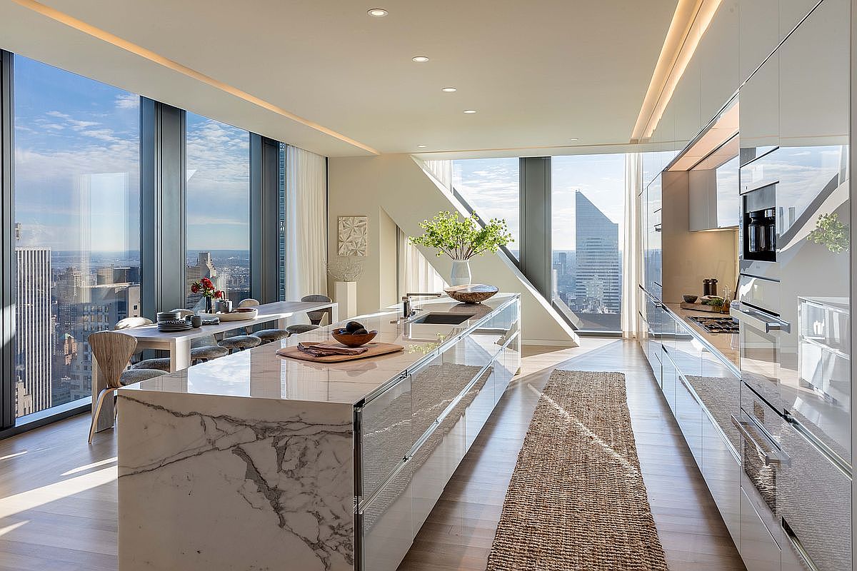 This ultra-modern, high-rise kitchen features a massive stone-topped island with integrated cabinetry and a sleek, minimalist aesthetic. The space is defined by expansive floor-to-ceiling windows that offer panoramic city views, flooding the room with natural light. A long dining table sits adjacent to the kitchen, creating a seamless open-concept layout perfect for sophisticated urban living.