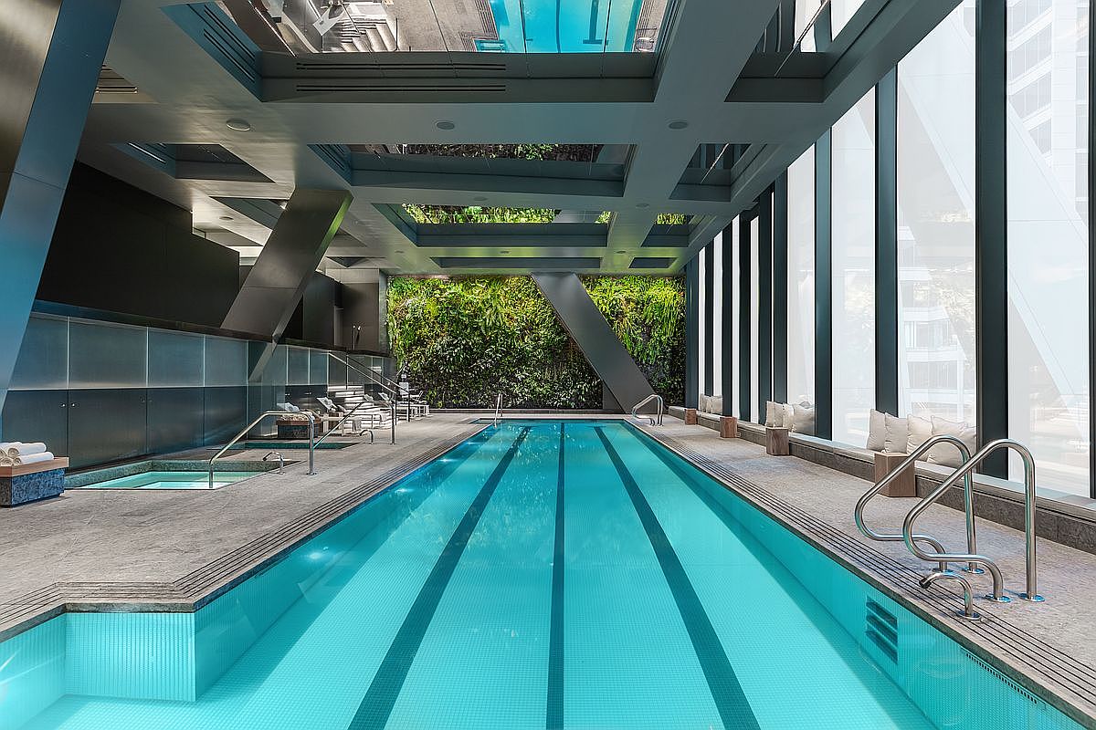 This sophisticated indoor lap pool area features a long, rectangular swimming pool with dark lane markers and a separate adjacent spa. The space is defined by a striking living green wall at the far end, complemented by modern architectural columns and large windows that provide ample natural light. The sleek, contemporary design is enhanced by a reflective ceiling structure and minimalist seating along the side, creating a serene and luxurious atmosphere.