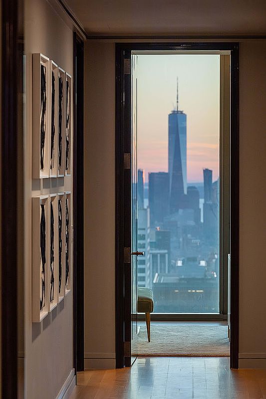 This interior view captures a sophisticated hallway leading toward a glass-paned doorway that frames a dramatic, elevated view of the New York City skyline, including the One World Trade Center. The hallway walls are adorned with a series of framed, abstract black-and-white artworks, creating a gallery-like atmosphere. The perspective draws the eye through the transition space, highlighting the contrast between the warm, refined interior and the expansive urban vista beyond.
