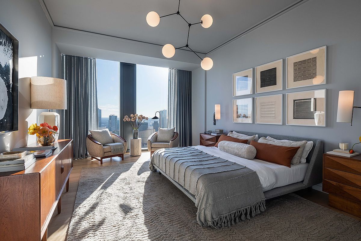 This sophisticated primary bedroom features a contemporary design with a neutral color palette and high-end finishes. A large bed with a textured gray throw sits center stage, flanked by wooden nightstands and illuminated by a modern, multi-globe chandelier. The room is anchored by floor-to-ceiling windows that offer expansive city views, while a gallery wall of framed art adds a curated, artistic touch to the space.