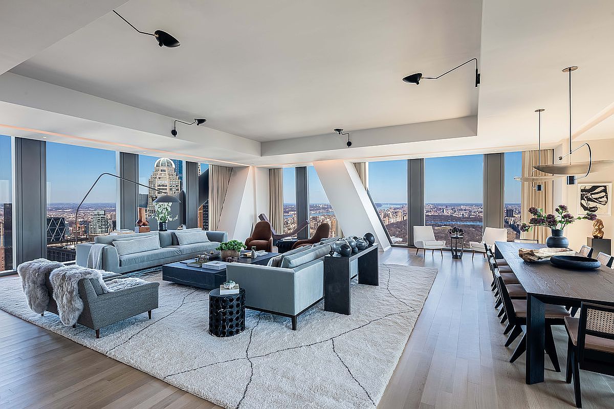 This expansive, open-concept living and dining area features a sophisticated, modern aesthetic with light wood flooring and a neutral color palette. The space is defined by massive floor-to-ceiling windows that provide panoramic city views, including a clear sightline to a large urban park. The room is furnished with plush, contemporary seating, a large area rug, and a long dining table, all illuminated by minimalist, sculptural lighting fixtures.
