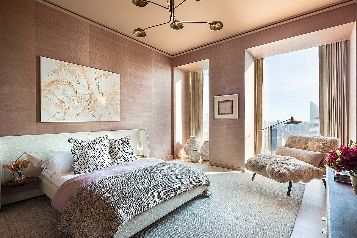 This sophisticated primary bedroom features a neutral color palette with textured wall coverings and a plush, upholstered bed frame. Large windows provide expansive city views and bathe the room in natural light, while a cozy, fur-covered lounge chair creates a serene reading nook. The space is accented by a modern chandelier, abstract wall art, and elegant bedside lighting, offering a luxurious and tranquil atmosphere.