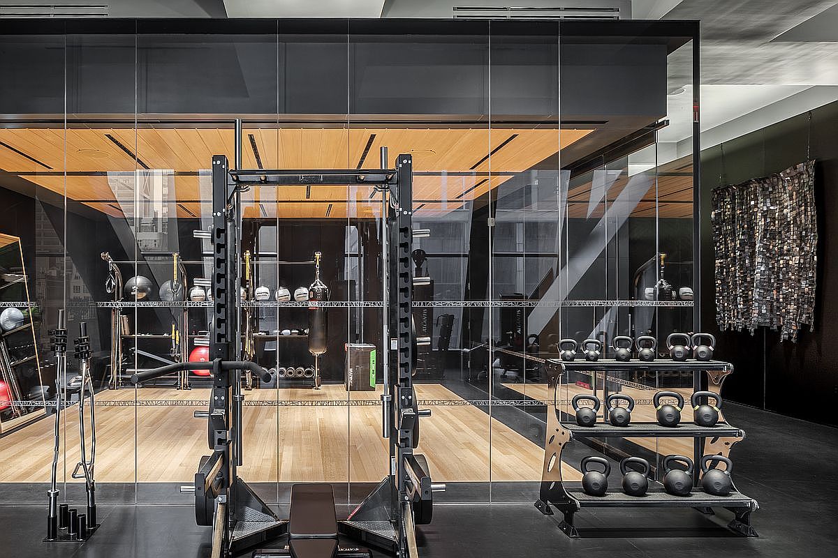 This sophisticated home gym features a central power rack and a dedicated rack of kettlebells, all set against a backdrop of light wood flooring and dark, modern accents. The space is enclosed by floor-to-ceiling glass walls, creating an open and airy atmosphere while maintaining a distinct workout zone. A unique, textured wall art piece hangs to the right, adding a touch of industrial-chic character to the high-end fitness environment.