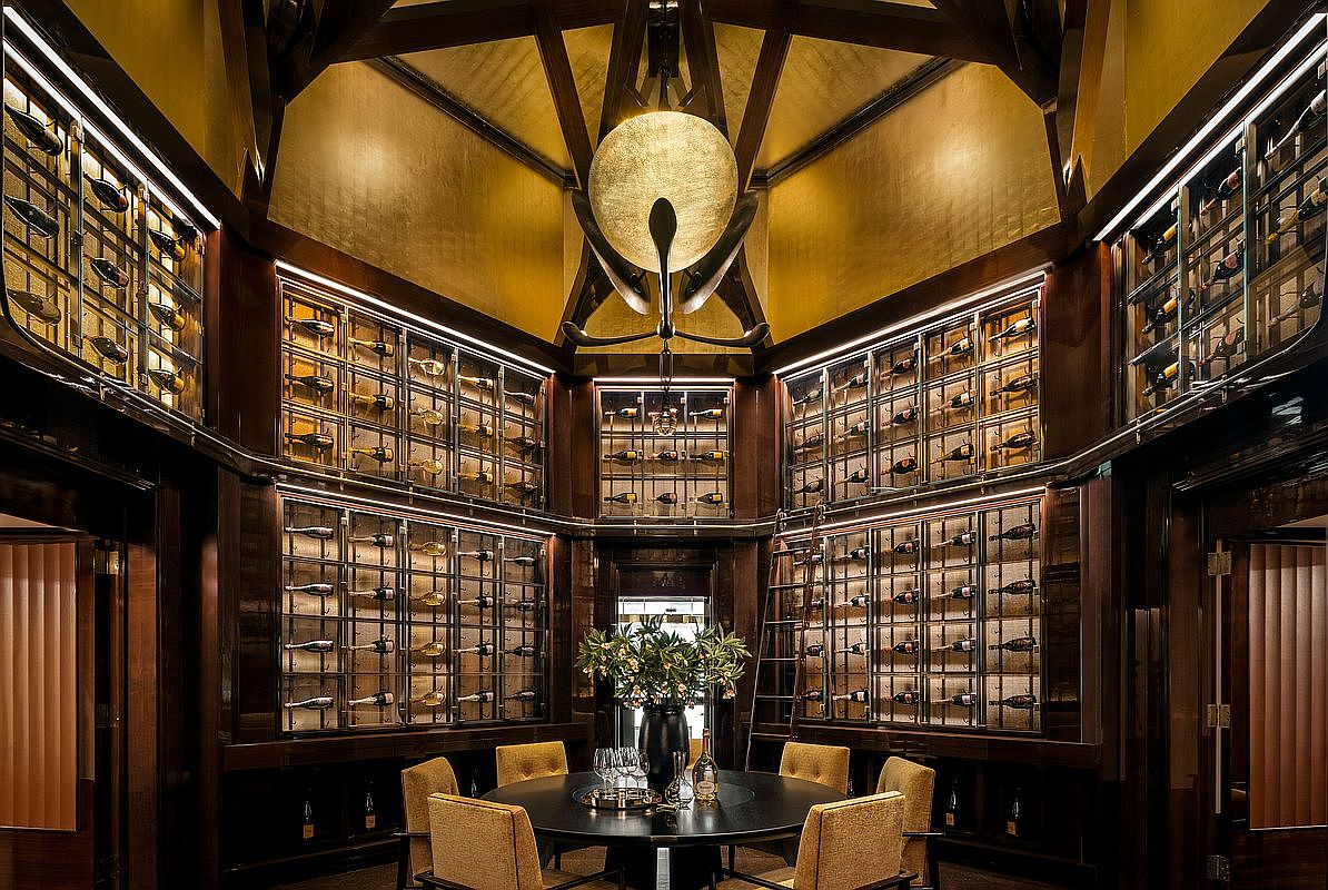 This sophisticated wine cellar features a central circular dining table surrounded by custom, dark wood cabinetry with glass-fronted storage for an extensive wine collection. The room is anchored by a dramatic, large circular light fixture suspended from a vaulted, gold-toned ceiling, creating an opulent and intimate atmosphere. The symmetrical layout and rich, warm wood tones provide a luxurious setting for private tastings and gatherings.