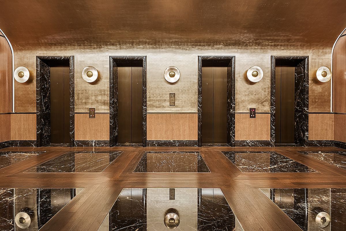 This sophisticated elevator lobby features a symmetrical layout with four dark metal elevator doors framed by polished black stone trim. The walls are finished in a metallic, textured material, accented by circular wall sconces and a central floor indicator panel. The floor is composed of a geometric pattern of dark stone tiles inlaid within a warm-toned wood border, creating a luxurious and formal atmosphere.