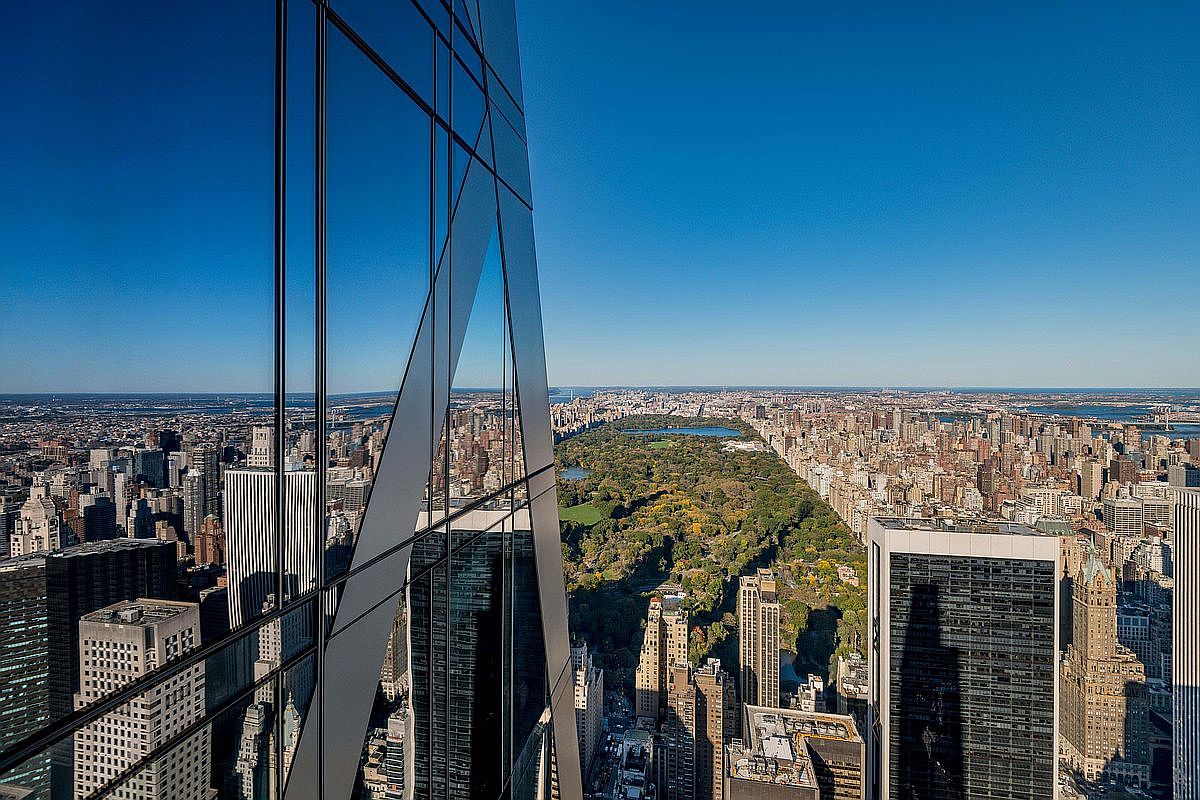 This high-altitude aerial perspective captures a dramatic view of Central Park in New York City, framed by the sharp, angular glass facade of a modern skyscraper. The composition emphasizes the contrast between the lush green expanse of the park and the dense, surrounding urban grid of high-rise buildings. The reflection on the building's exterior glass adds a sophisticated, architectural layer to the expansive cityscape under a clear blue sky.
