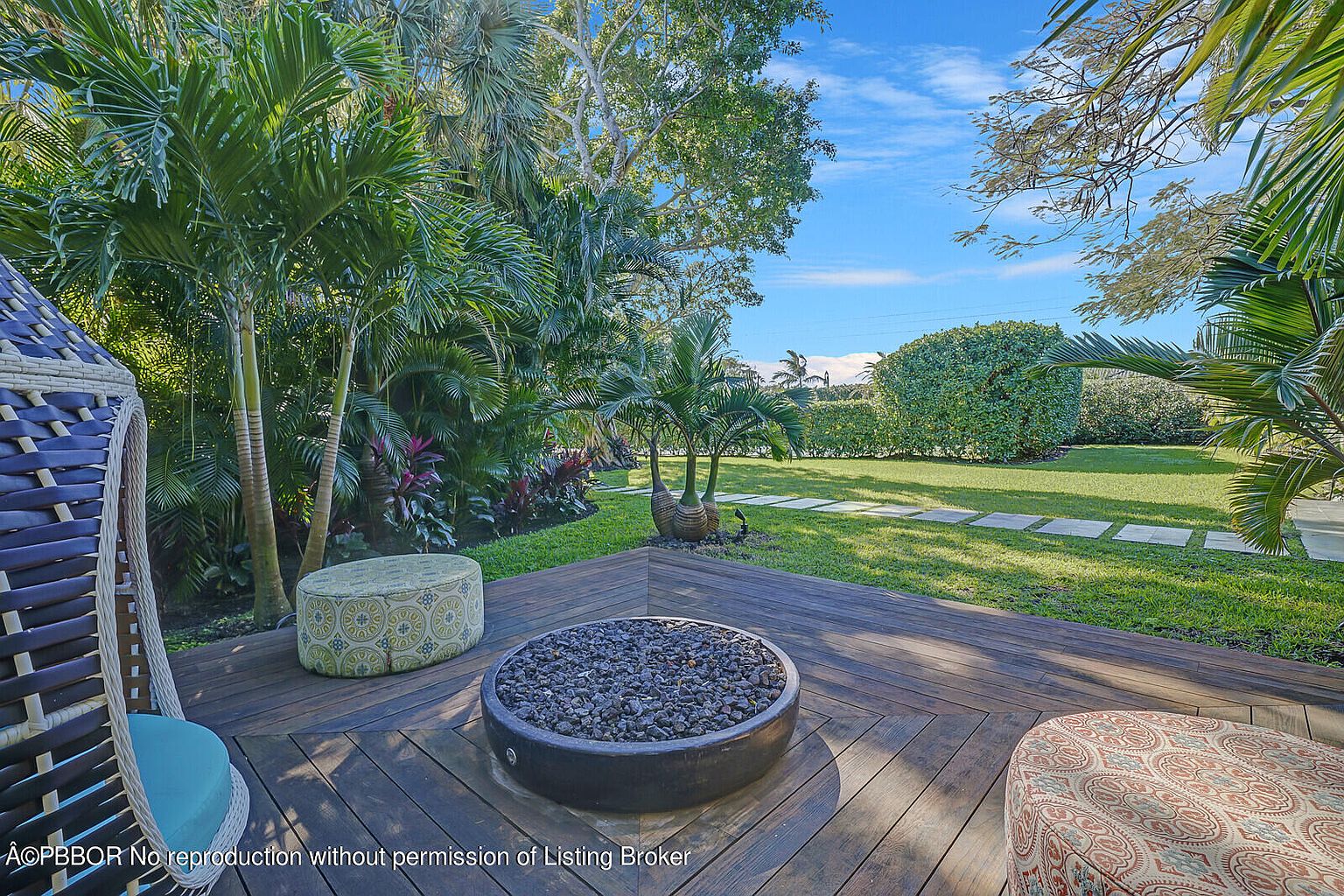 This image showcases a beautifully designed outdoor patio area, featuring a wooden deck with a central fire pit surrounded by comfortable seating. Lush tropical landscaping and manicured lawns create a serene and inviting atmosphere. The space is perfect for relaxation and entertaining, offering a seamless blend of nature and luxury.