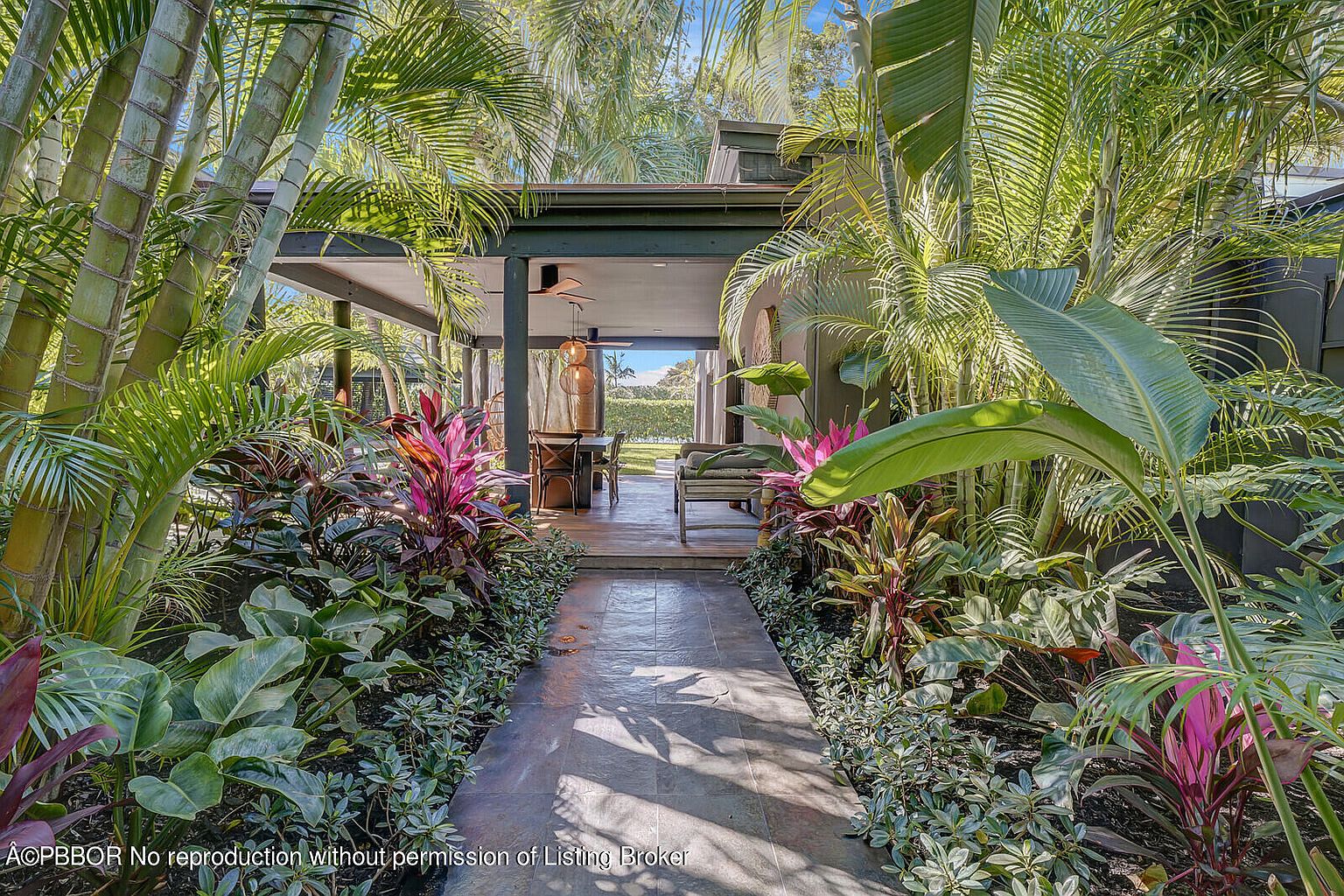 This image showcases a lush garden leading to an outdoor living space. The pathway is lined with vibrant tropical plants, creating a serene and inviting atmosphere. The covered patio features comfortable seating and a dining area, perfect for outdoor entertaining.