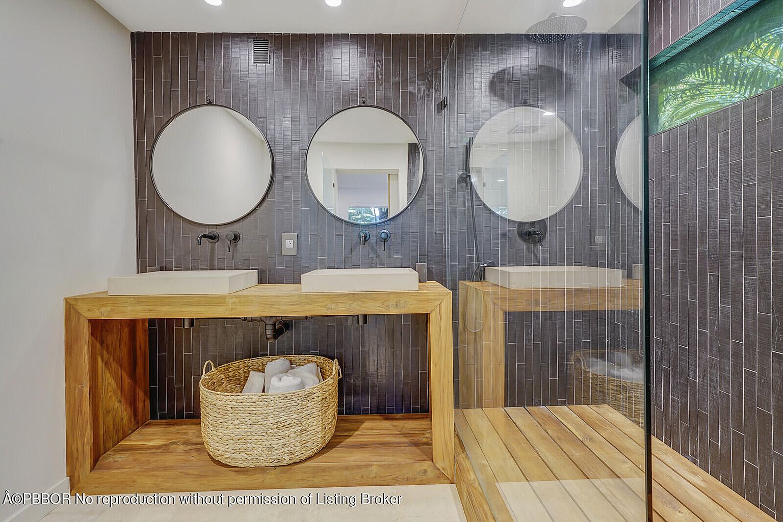 This is a modern primary bathroom featuring a double vanity with vessel sinks and round mirrors against a dark tiled wall. The vanity is made of light wood, and a woven basket sits underneath for storage. A glass-enclosed shower is visible on the right, showcasing a continuation of the dark tile and wooden flooring, creating a spa-like atmosphere.