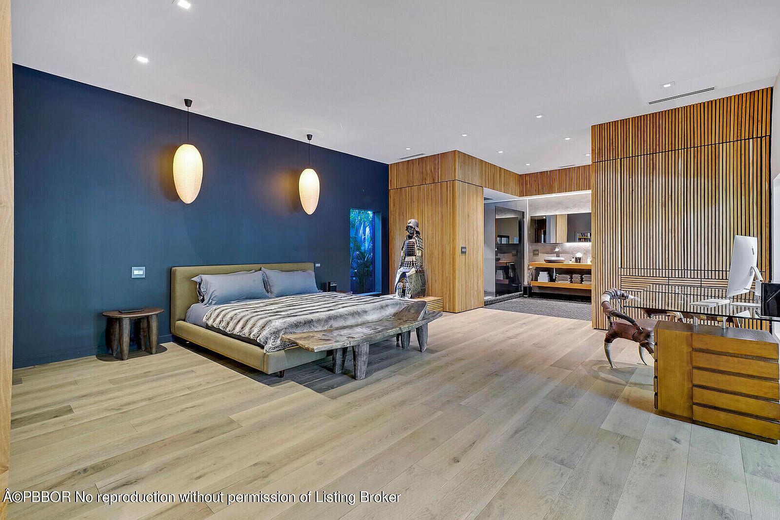 This is a spacious primary bedroom featuring a king-size bed with a unique wooden bench at its foot. The room has a modern aesthetic with a dark blue accent wall, pendant lighting, and light wood flooring. A desk area with a modern chair and computer is visible, along with a glimpse into an adjacent bathroom, creating a luxurious and functional space.