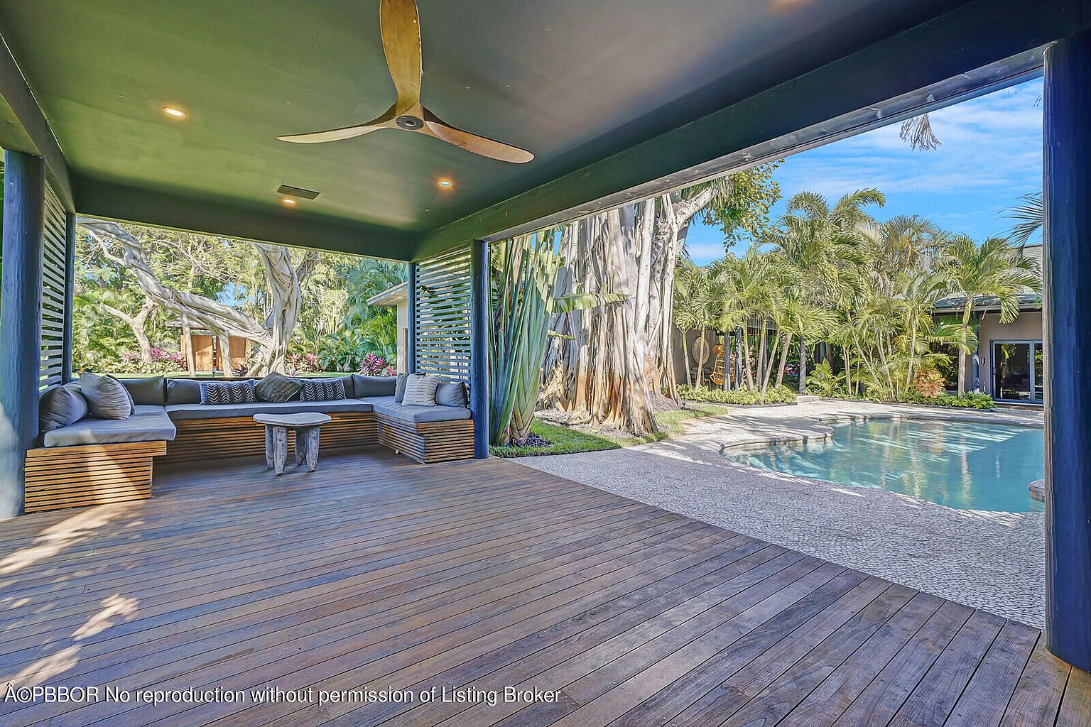 This image showcases a covered outdoor patio or deck area, featuring a built-in wooden seating arrangement with comfortable cushions. The space is open to a backyard with a swimming pool and lush tropical landscaping, creating a seamless indoor-outdoor living experience. A wooden ceiling fan adds to the relaxed and luxurious ambiance.