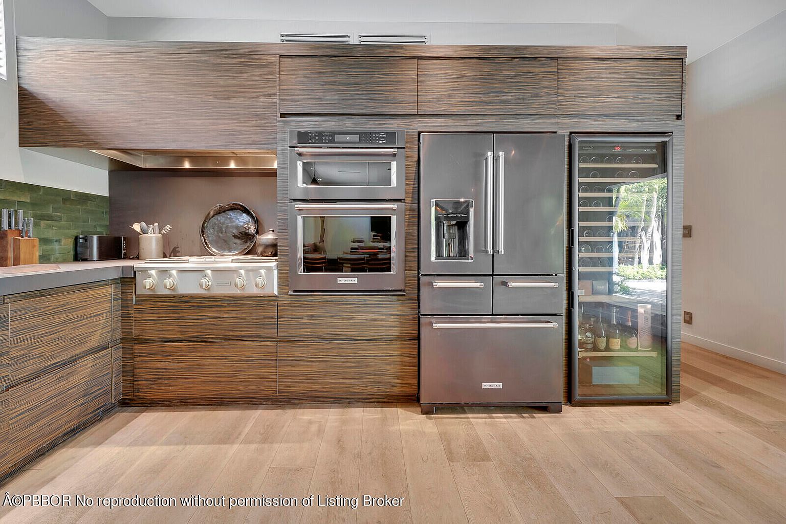This is a modern kitchen featuring dark wood-grain cabinetry and stainless steel appliances. The kitchen includes a built-in oven and microwave, a large refrigerator, and a wine cooler. The flooring is light wood, and the overall design is sleek and contemporary.