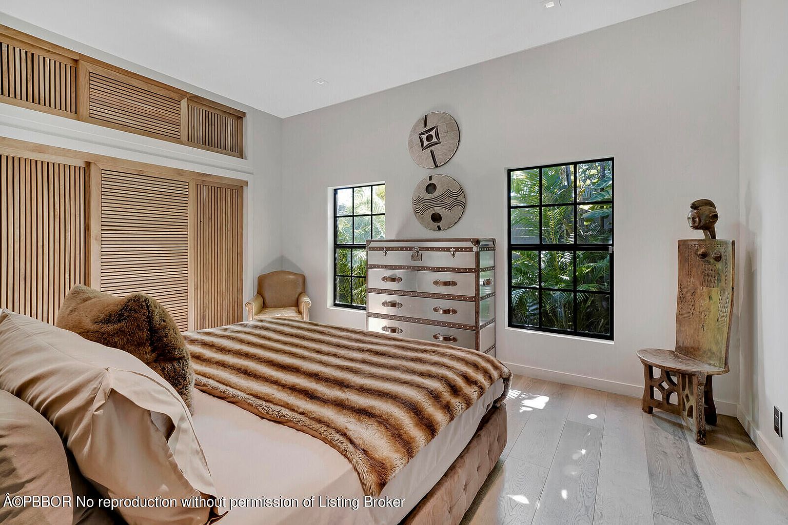 This is a primary bedroom featuring a bed with a striped throw, a silver dresser, and two windows that provide natural light. The room has a modern, minimalist style with light wood floors and white walls. There is also a unique wooden chair and statue in the corner, adding an artistic touch.