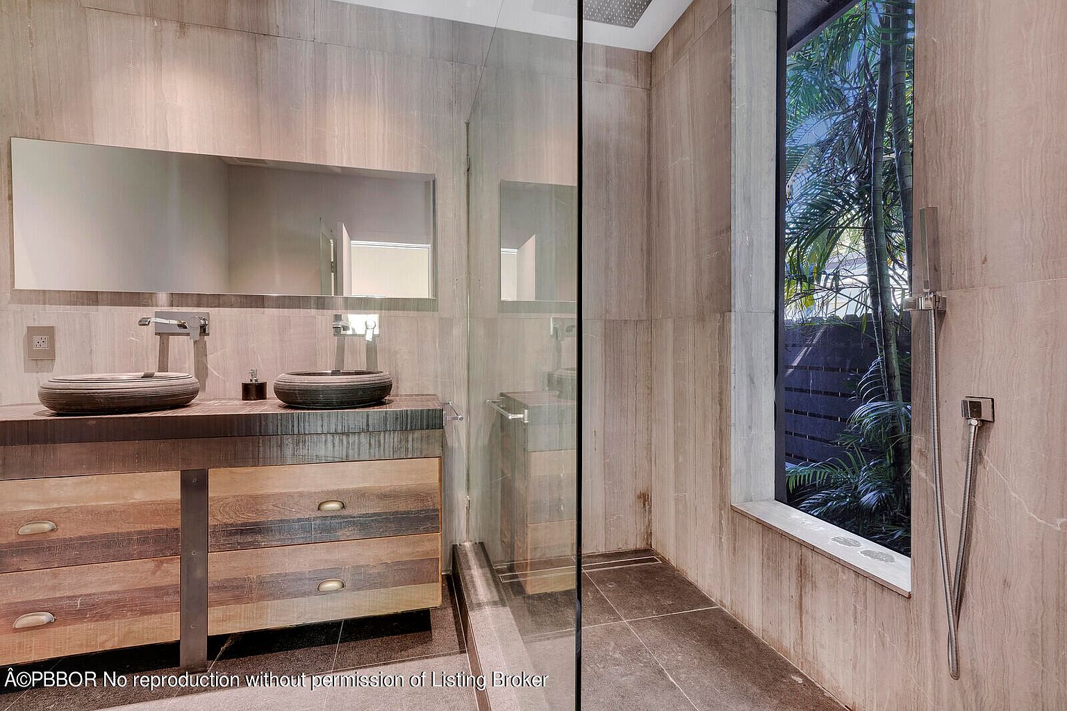 This is a modern primary bathroom featuring a double vanity with unique vessel sinks and a large mirror. The shower area is enclosed with glass and includes a window offering a view of lush greenery. The walls and floors are tiled in a neutral tone, creating a spa-like atmosphere.