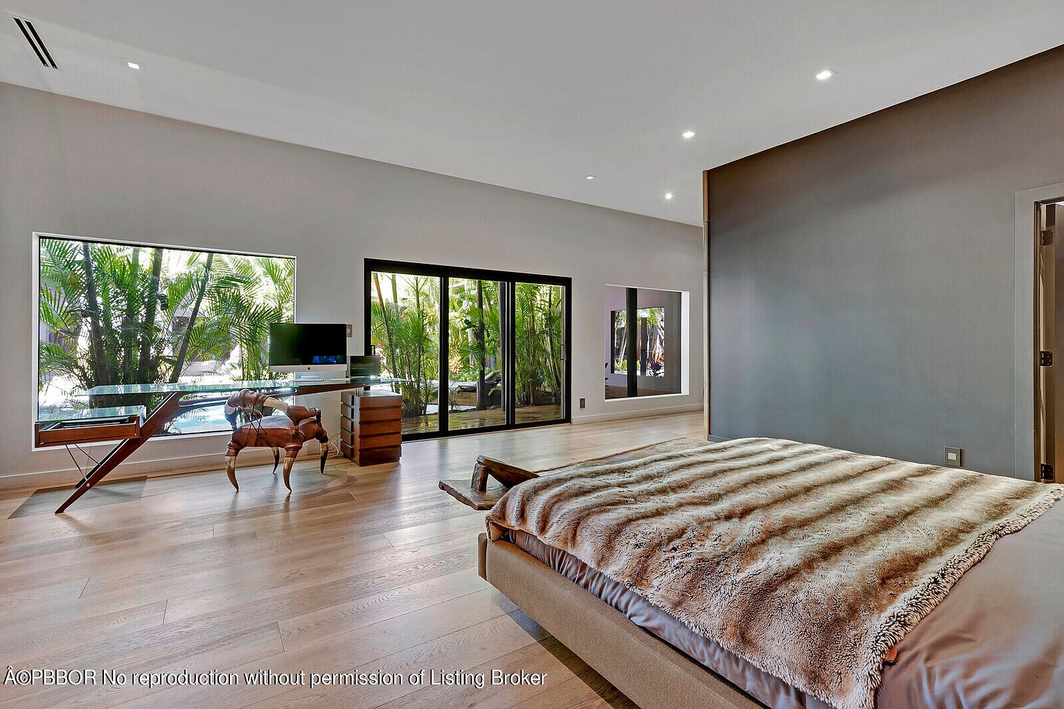 This is a spacious primary bedroom featuring a modern design with clean lines and natural light. A large window and sliding glass door offer views of lush greenery, while a unique glass-top desk with wooden accents adds character. The bed is covered with a plush, striped throw, and the hardwood floors contribute to the room's warm and inviting atmosphere.