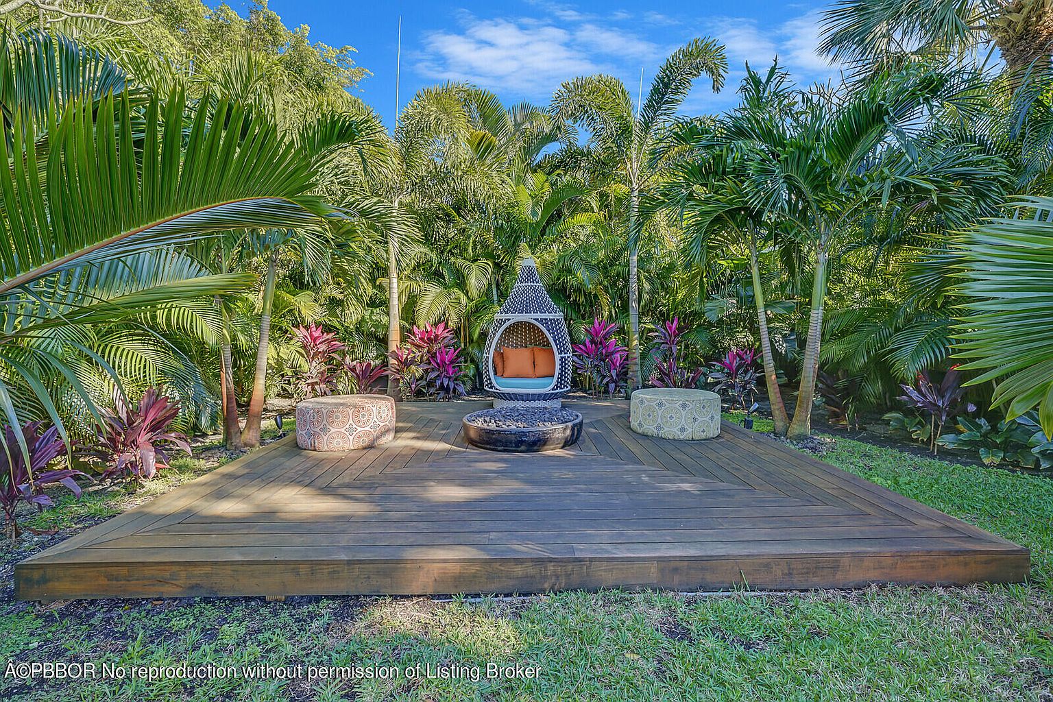 This image showcases a beautifully landscaped backyard featuring a wooden deck with a unique seating area. The focal point is a stylish hanging chair and a modern fire pit, surrounded by lush tropical plants and palm trees. The scene evokes a sense of relaxation and outdoor living, perfect for entertaining or enjoying a peaceful retreat.