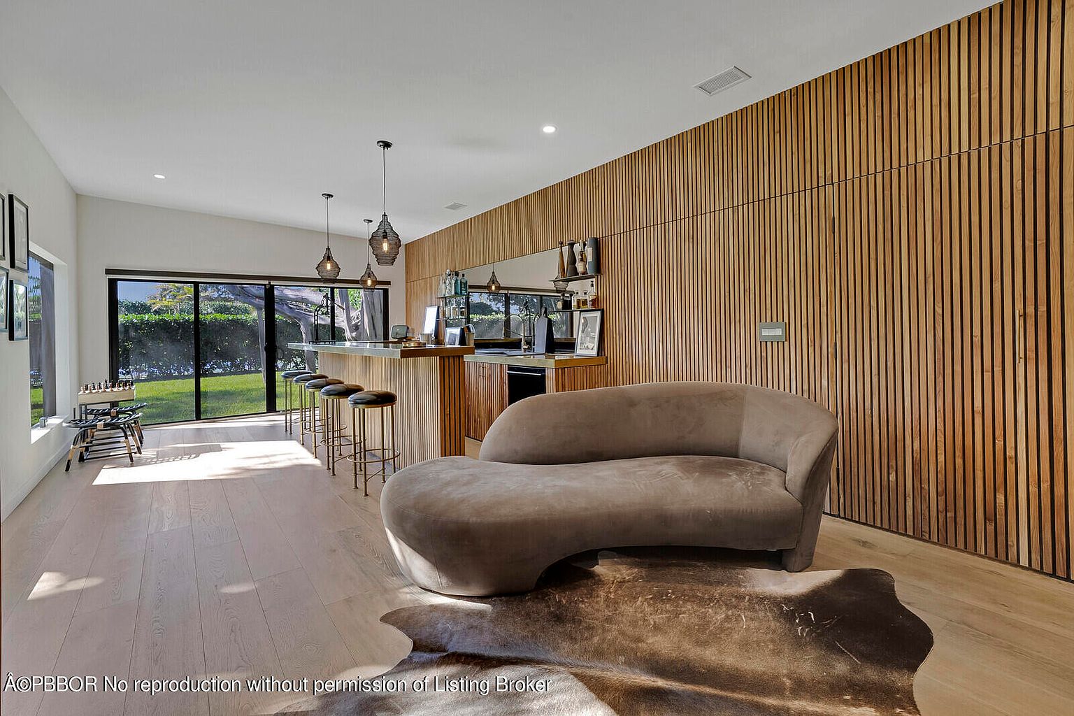 This interior shot showcases a modern living room with a unique design. A curved, brown sofa sits on a cowhide rug, complemented by a wooden slat wall and a built-in bar area with pendant lighting. The room features light wood flooring and a large window offering natural light, creating a stylish and inviting atmosphere.