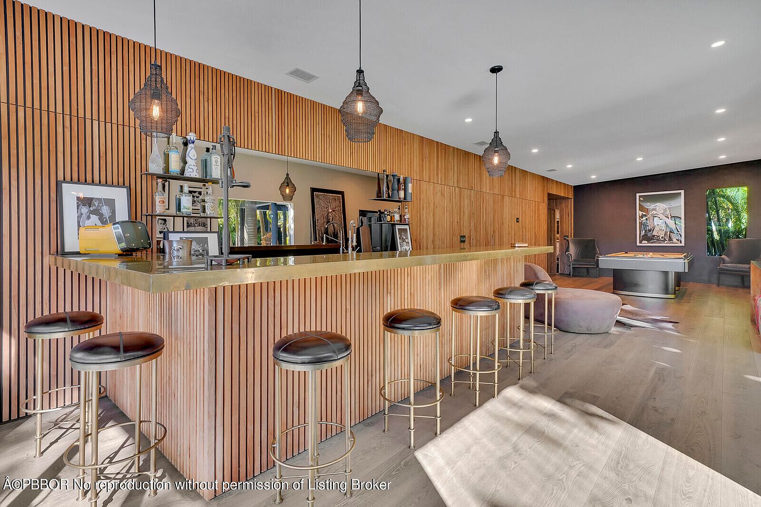 This interior shot showcases a stylish living room with a modern bar area. The bar features a unique wooden facade and a brass countertop, complemented by several bar stools. The room also includes a pool table and comfortable seating, creating an inviting and sophisticated entertainment space.