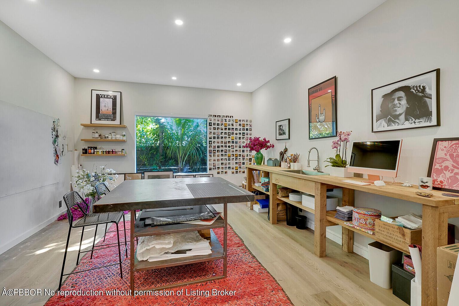 The image showcases a well-lit and organized office/study space. A large work table with metal legs and a dark top sits on a red patterned rug, complemented by a long wooden counter with a sink and an iMac computer. The room features a collection of artwork and photographs on the walls, creating an inspiring and creative atmosphere.