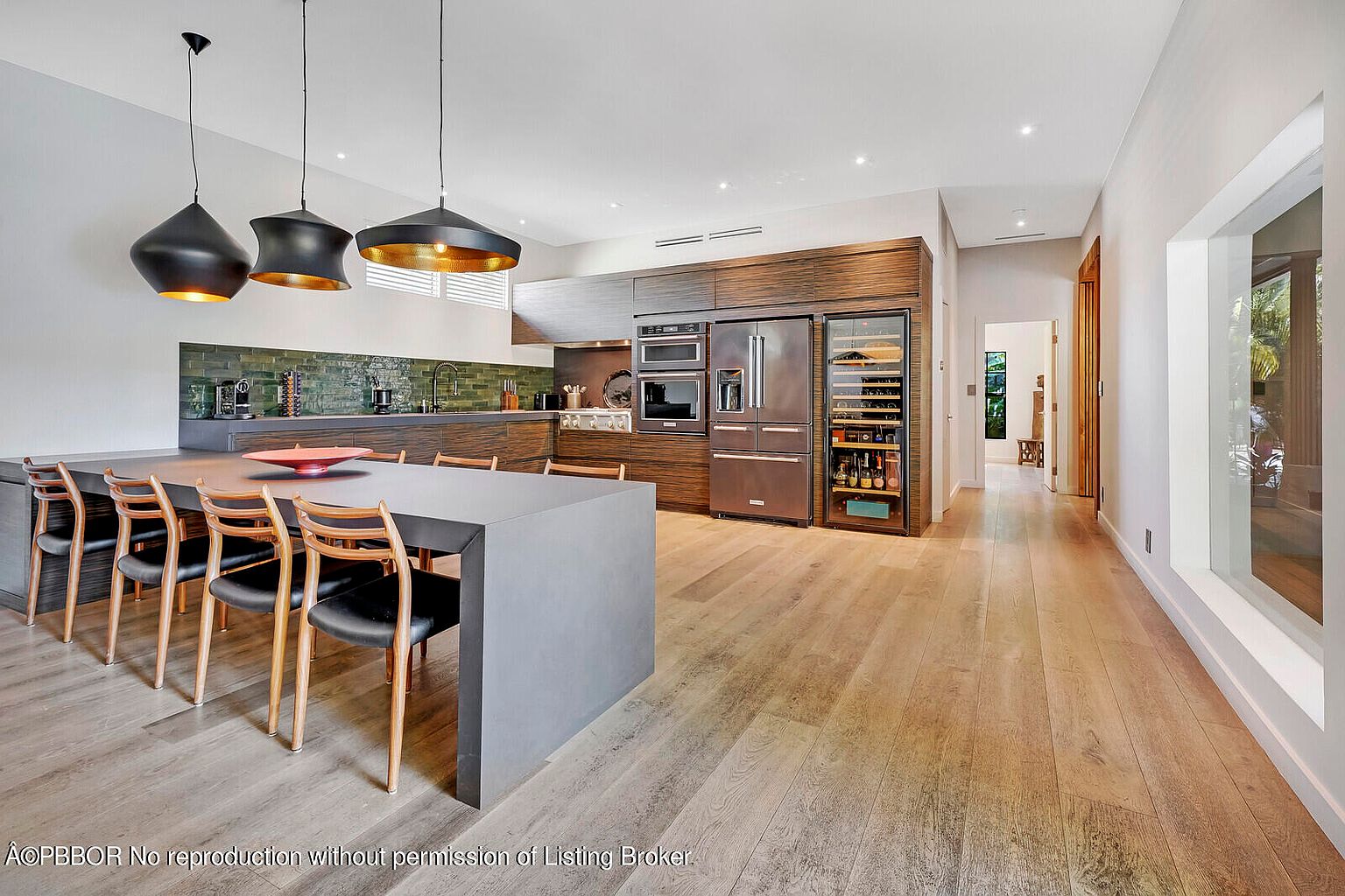 This is a modern kitchen with dark wood cabinetry, stainless steel appliances, and a large gray island with seating. Three pendant lights hang above the island, and a green tile backsplash adds a pop of color. The kitchen has an open floor plan and hardwood floors, creating a sleek and inviting space.