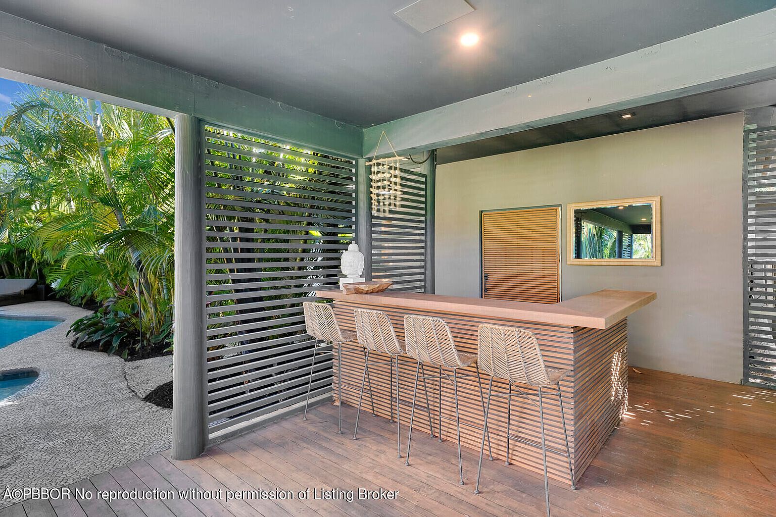 This image showcases an outdoor bar area on a patio or deck. The bar features a wooden counter and base with horizontal slats, complemented by four woven bar stools. The space is partially enclosed by slatted screens, offering privacy while allowing natural light and views of the surrounding greenery and pool area.