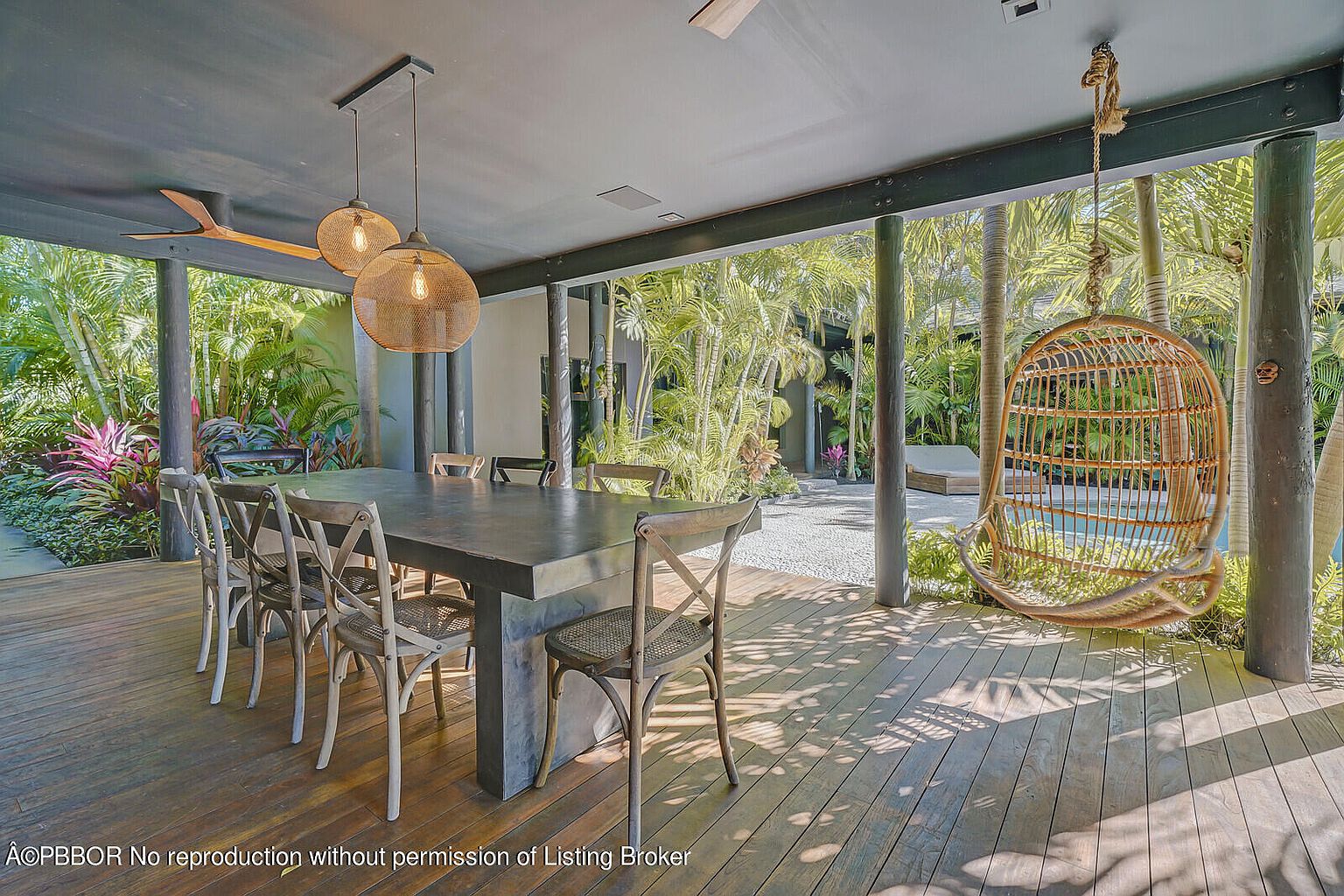 This image showcases a covered outdoor dining and relaxation area, featuring a large wooden table with chairs, pendant lighting, and a hanging wicker chair. The space opens to a lush tropical garden with a glimpse of a pool, creating a serene and inviting atmosphere. The wooden deck and dark ceiling add a touch of rustic elegance.