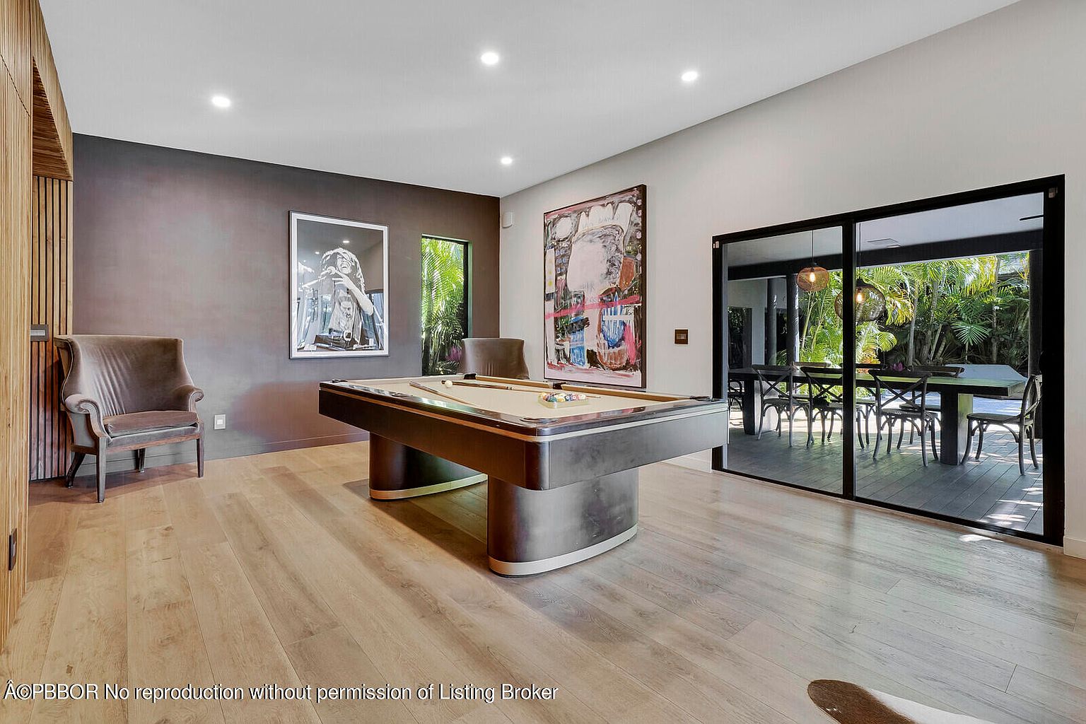 This interior shot showcases a stylish living room featuring a pool table as a central element. The room is decorated with modern art and has a sophisticated color palette, complemented by hardwood flooring. Large sliding glass doors provide access to an outdoor dining area and lush greenery, enhancing the indoor-outdoor flow.