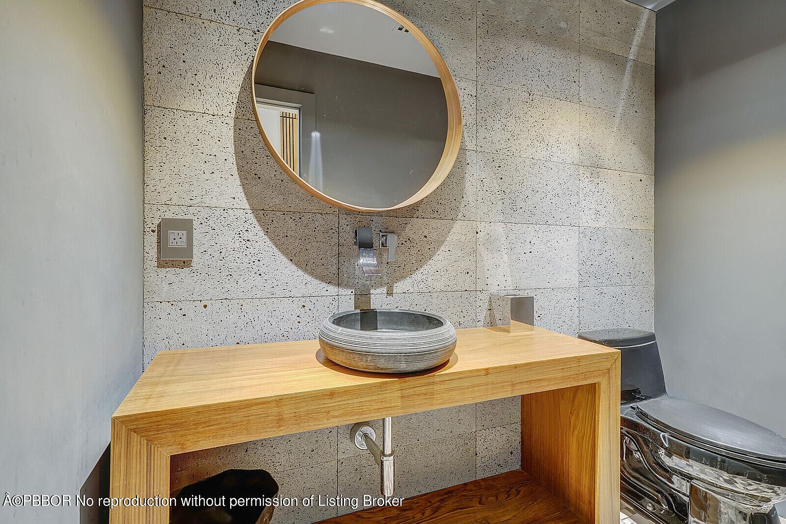 This is a modern guest bathroom featuring a round mirror above a unique vessel sink on a wooden vanity. The walls are covered in textured gray tiles, and a sleek, black toilet is visible to the right. The design is minimalist and stylish, creating a sophisticated and clean space.