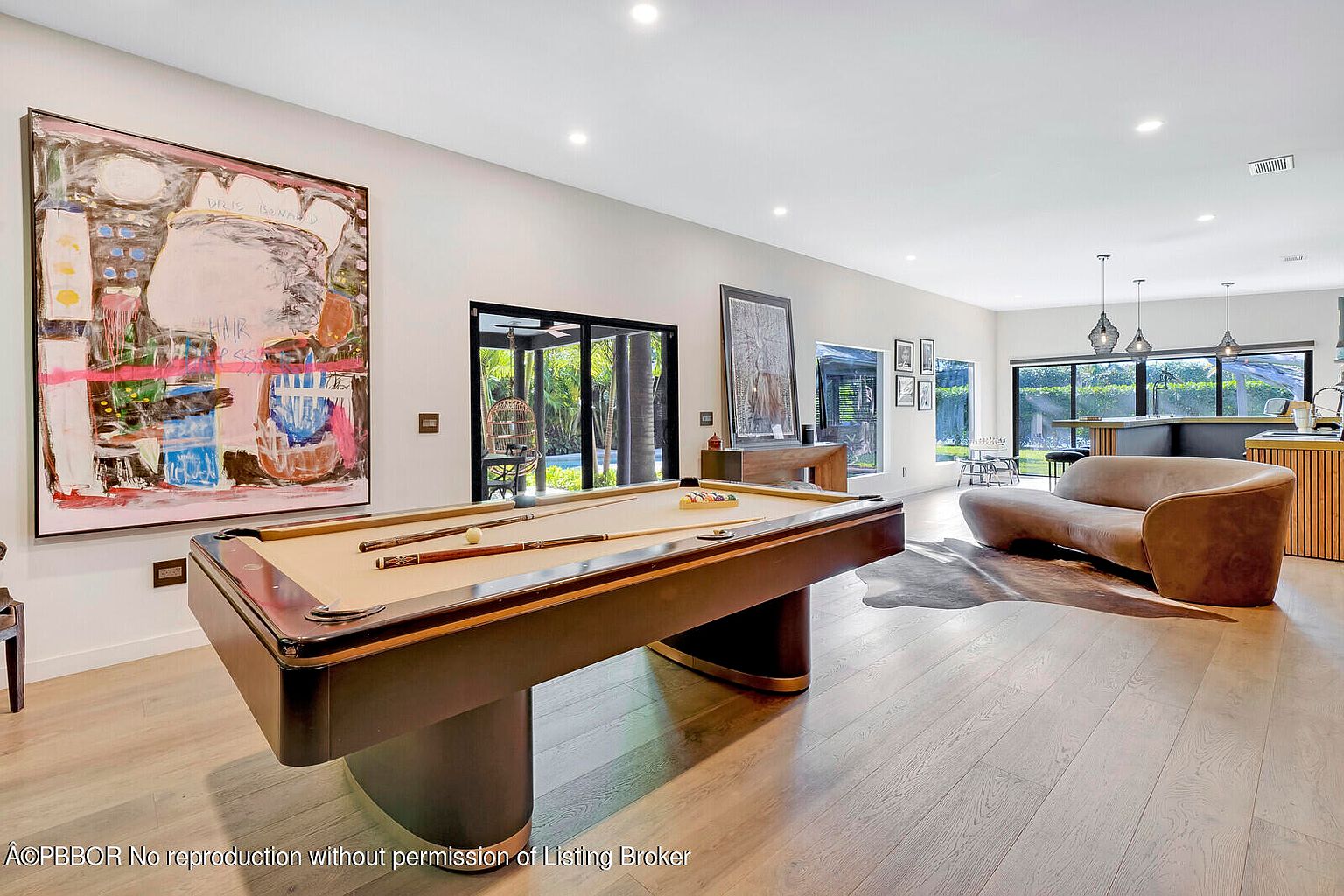 This interior shot showcases a spacious living room featuring a billiard table as a central element. The room is decorated with modern art and includes a unique curved sofa, complemented by light hardwood flooring. Large windows and sliding glass doors provide ample natural light and views of the exterior, enhancing the open and airy feel of the space.