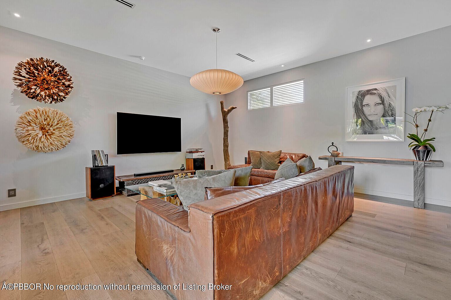 This is an interior shot of a living room featuring a large, brown leather sectional sofa as the focal point. The room is decorated with modern art pieces, including two decorative wall hangings and a framed portrait. The flooring is light-colored wood, and a unique pendant light hangs from the ceiling, creating a warm and inviting atmosphere.