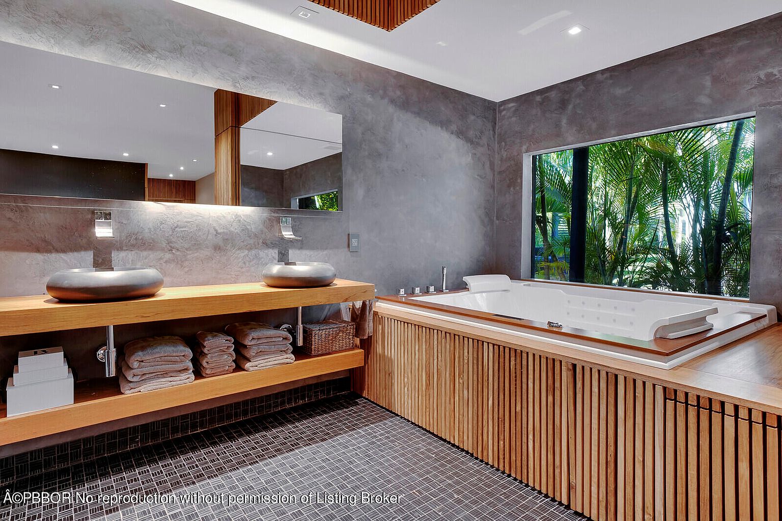 This is a luxurious primary bathroom featuring a modern design with gray textured walls and a unique wooden vanity with dual vessel sinks. A large soaking tub is positioned by a window offering a view of lush greenery, creating a spa-like atmosphere. The floor is tiled with a dark, geometric pattern, adding to the contemporary aesthetic.