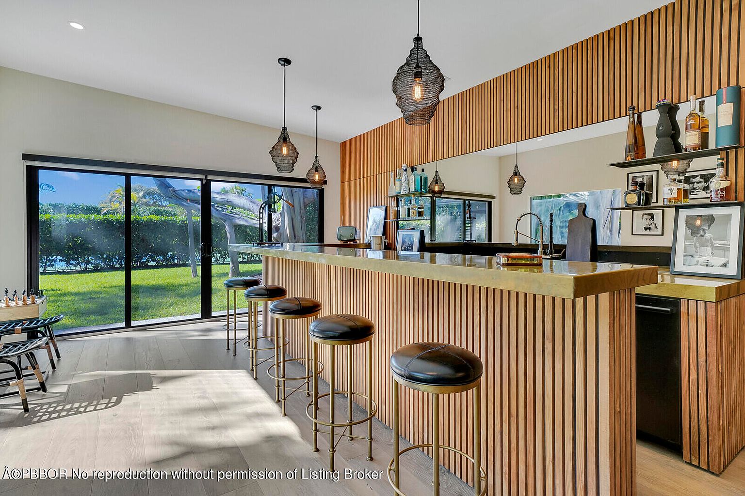 This interior shot showcases a stylish bar area within a living space, featuring a wood-paneled bar with a brass countertop and matching bar stools. Pendant lighting hangs above the bar, and large windows offer a view of the outdoor greenery, creating a sophisticated and inviting atmosphere. The room is well-lit, highlighting the modern design and high-end finishes.