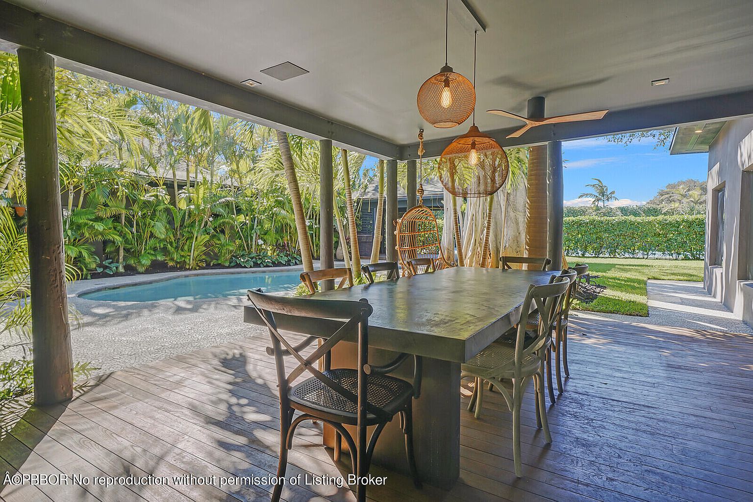 This image showcases a covered outdoor dining area with a large concrete table and various chairs, situated on a wooden deck. The space overlooks a lush tropical garden with a swimming pool, creating a serene and inviting atmosphere. Hanging pendant lights add a touch of elegance, while the open design seamlessly blends indoor and outdoor living.